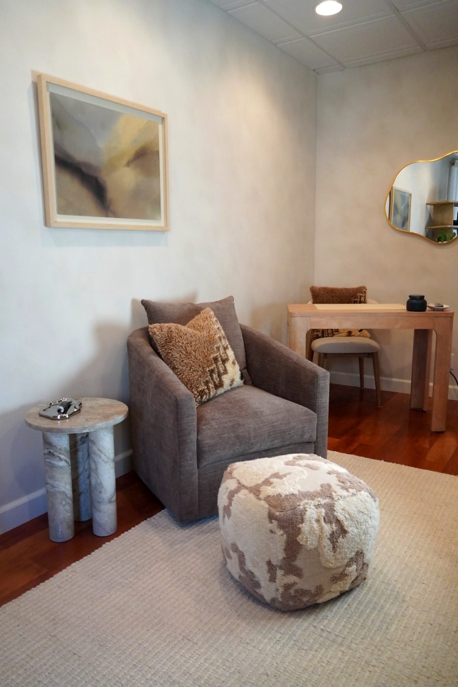 Reading nook with a swivel armchair, marble side table, sheepskin pouf, and a small writing desk under a wavy gold mirror.